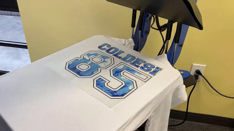 A Heat Press Machine Applying a Design with Large Text and Numbers onto A White Shirt