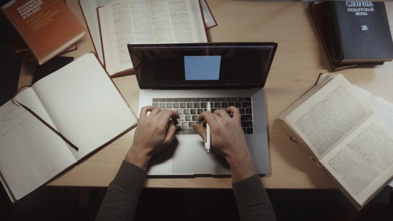 A person typing on a laptop surrounded by books and notebooks, working on long-form educational content