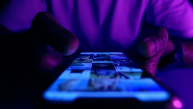 Close-up of a person using a smartphone under purple lighting, browsing a grid of images on the screen