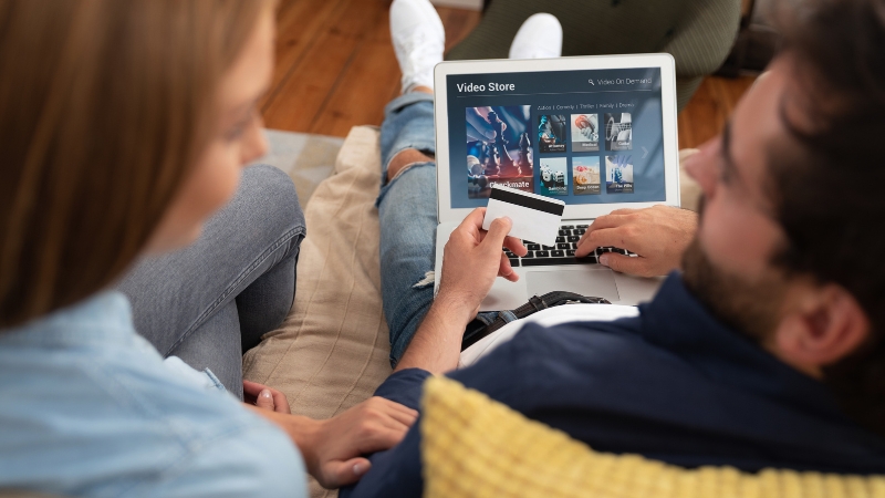 A couple sits on a couch holding a credit card while choosing a video subscription service on a laptop