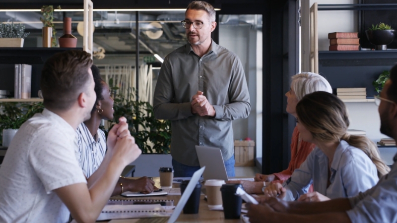 A group of colleagues sits at a table as one person stands and leads a discussion
